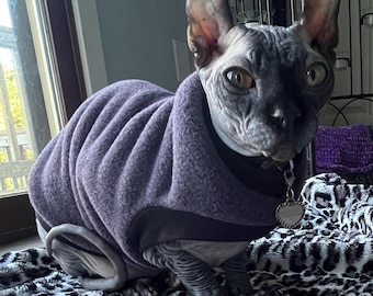 May include: A hairless cat wearing a purple fleece sweater with a black trim. The cat is sitting on a black and white patterned blanket. The cat has a silver heart-shaped tag on a silver chain around its neck.