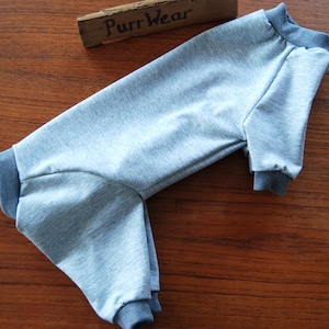 Op de afbeelding: Een grijze onesie voor huisdieren met donkergrijze manchetten en kraag. De onesie heeft lange mouwen en pijpen. De tekst "Purr Wear" staat op een houten bord op de achtergrond.