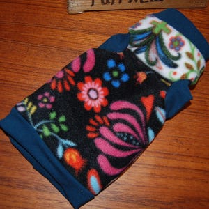 May include: A fleece pet sweater featuring a black base with a vibrant floral pattern in shades of pink, orange, blue, and green. The garment includes solid dark blue trim at the neck and arm openings, with a white-based floral collar accent.