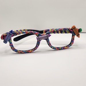 May include: Spectacles with black frames, decorated with colourful rubber bands. Small crayon-shaped embellishments are attached to the frames, each labelled "CRAYON". The rubber bands are woven around the frames in a rainbow pattern.