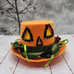 May include: Orange felt top hat with a jack-o'-lantern face design. The hat features black gem accents for eyes and mouth, a green ribbon band, and a small cluster of berries. The hat sits on a matching orange brim.
