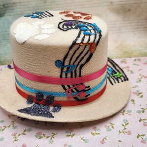 Puede incluir: Un sombrero de copa beige con notas musicales y huellas de pata. El sombrero presenta una cinta rosa y azul. Las notas musicales y la huella de pata están adornadas con purpurina y lentejuelas.