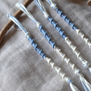 May include: Four handmade friendship bracelets with blue and white twisted thread. The bracelets are arranged diagonally on a neutral linen surface, with a wooden element in the background. Each bracelet is a simple, elegant accessory.