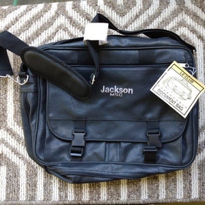 May include: Black leather document bag with a shoulder strap and a buckle closure. The bag has a label that says "Jackson MSC" and another label that says "Le Club Expandable Document Bag".