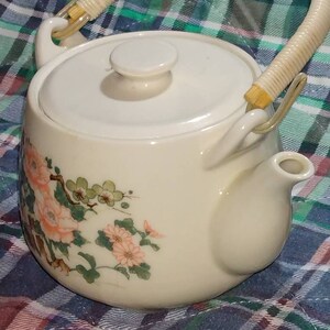 May include: A white ceramic teapot with a floral design and a woven handle. The teapot has a lid and a spout. The teapot is sitting on a plaid tablecloth.
