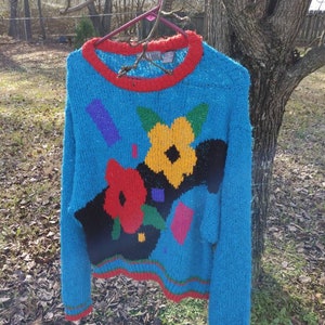 May include: A blue fuzzy sweater with a colorful abstract floral design. The sweater has a red, green, and black trim.