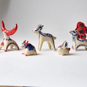 Peut inclure: Une collection de sept petites figurines d'animaux en céramique de couleur crème, bleue et rouge. L'ensemble comprend des créatures stylisées avec des motifs peints, dont une figure à cornes, un cerf et un lapin, aux surfaces lisses.