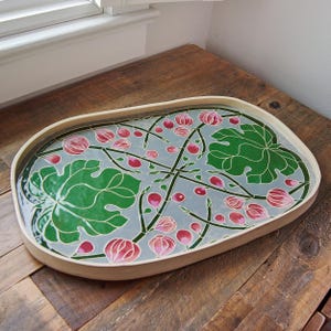 May include: An oval-shaped decorative tray with a light gray background and a botanical design. The design features green leaves, pink flower buds, and stems. The tray has a light brown wooden frame.