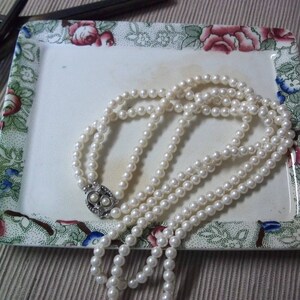 May include: A three-strand pearl necklace with a decorative clasp rests on a rectangular, floral-patterned dish. The dish features a white center and a border with red, pink, blue, and green floral designs. Jewelry and vintage theme.