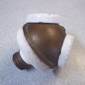 May include: A vintage bottle stopper with a brown metal body and white ceramic accents. The stopper has a rounded shape with a cylindrical base and a circular top. The white ceramic sections are slightly textured.