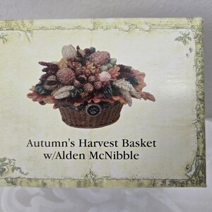 May include: A decorative box featuring a detailed illustration of a woven basket overflowing with autumn harvest elements. The basket contains pinecones, leaves, and flowers in shades of brown, pink, and green. The text "Autumn's Harvest Basket w/Alden McNibble" is below the basket.