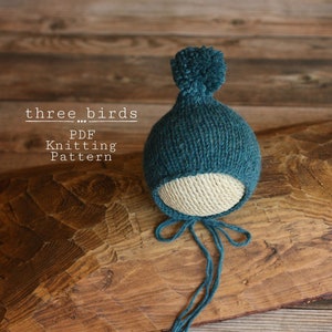 Puede incluir: Un gorro de bebé de punto azul turquesa con un borde blanco y un pompón en la parte superior. El gorro está atado con una cinta azul turquesa. El gorro está sobre una superficie de madera. El texto "three...birds PDF Knitting Pattern" es visible en la imagen.