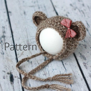 Könnte beinhalten: Gehäkelter Babyhut in Braun in Form eines Bären, mit kleinen Ohren und einer rot-weiß karierten Schleife. Der Hut hat lange geflochtene Bänder. Der Text "PDF Pattern" ist sichtbar.