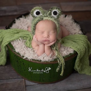 Könnte beinhalten: Ein Neugeborenes trägt eine grüne Froschkappe mit großen weißen Augen und liegt in einem grünen Eimer mit einer weißen, flauschigen Auskleidung. Das Baby schläft mit den Händen unter dem Kinn.