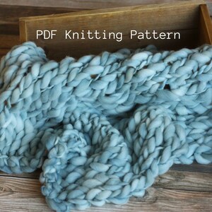 May include: A light blue knitted blanket with a chunky knit texture. The blanket is folded and draped over a wooden box with the text "PDF Knitting Pattern" printed on the box.