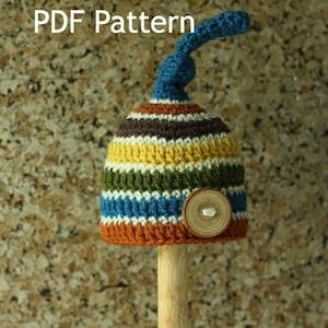 Peut inclure: Un bonnet en crochet rayé avec un dessus bleu et un bouton en bois. Le bonnet est présenté sur un bâtonnet en bois. Le texte "PDF Pattern" est visible en haut de l'image.