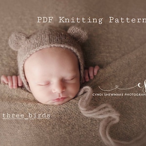 May include: A PDF knitting pattern for a brown knitted baby hat with bear ears. The hat is on a brown blanket with the text "three...birds" and "CYNDI SHEWMAKE PHOTOGRAPHY" visible.