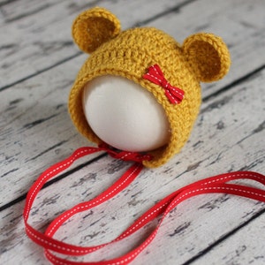 Puede incluir: Un gorro de bebé de crochet amarillo con orejas de oso y un lazo rojo. El gorro tiene un lazo de cinta rojo.