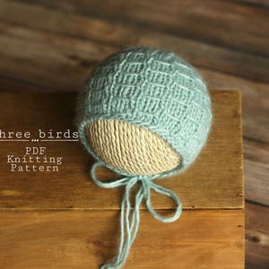 May include: A light blue knitted baby hat with a white lining and a tie. The hat is on a wooden surface. The text "three...birds PDF Knitting Pattern" is visible on the surface.