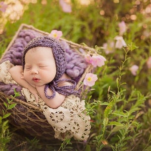May include: A sleeping baby wearing a purple knitted hat and a white crocheted blanket, lying in a woven basket filled with purple yarn. The basket is surrounded by green grass and pink flowers.