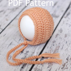 Könnte beinhalten: Eine babyfarbene gehäkelte Baby-Mütze mit langen Bändern. Die Mütze sitzt auf einem weißen Styropor-Kopf und die Bänder liegen auf einer hellbraunen Holzoberfläche. PDF-Muster.