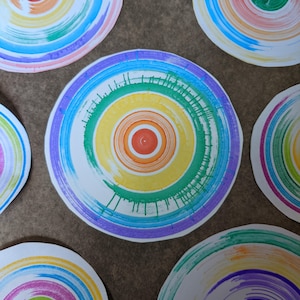 May include: Five colourful circular paper plates with concentric circles of different colours, including red, orange, yellow, green, blue, and purple. The plates are arranged on a brown surface.
