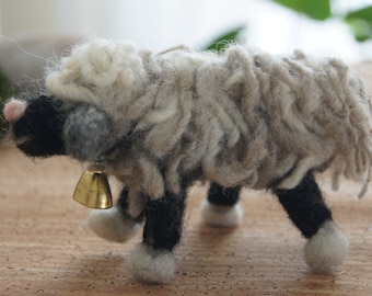 May include: A small, white and gray wool sheep with black legs and a gold bell around its neck. The sheep is standing on a brown surface.