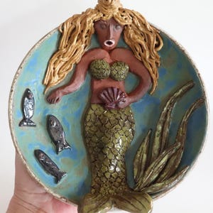 May include: A ceramic mermaid sculpture in a shallow bowl. The mermaid has brown skin, green scales, and long, golden hair. The bowl is blue with three black fish and green seaweed.