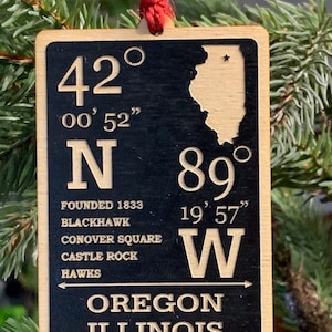 May include: Wooden Christmas ornament with a black background and tan border. The ornament features the text "OREGON ILLINOIS" and coordinates. The state of Illinois is also depicted. The ornament is hanging from a red string.