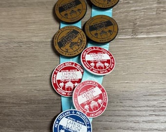 May include: A light blue lanyard with several round cruise ship medallions. The medallions are brown, red, and blue with text and images of cruise ships and destinations. The text includes "Carnival" and various cruise names.