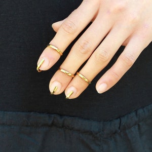 Puede incluir: Una mano que muestra anillos de u&ntilde;as y anillos de dedo dorados. Los anillos son finas bandas de metal. Los anillos de u&ntilde;as est&aacute;n curvados para adaptarse a la forma de la u&ntilde;a. El fondo es una camisa negra de manga larga.