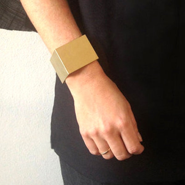 Könnte beinhalten: Ein goldfarbenes Metallarmband mit quadratischer Form. Das Armband ist an einer Seite offen und hat eine gebürstete Oberfläche.