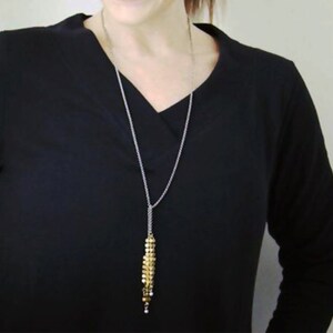 Puede incluir: Un collar largo de cadena dorada con un colgante de borla dorada. El collar lo lleva una persona con el pelo oscuro y una camisa negra.