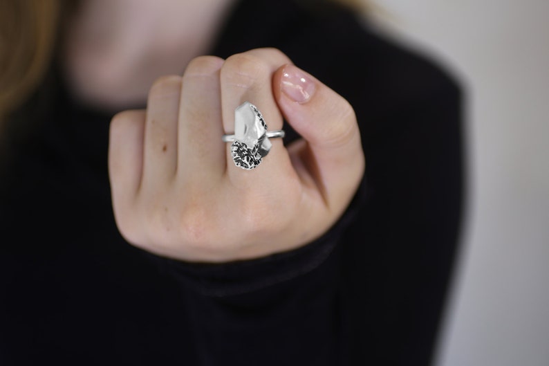 May include: A silver ring featuring a large, irregular-shaped, clear stone with a dark, textured base. The ring is held up against a black background, showcasing the jewelry's design and details.