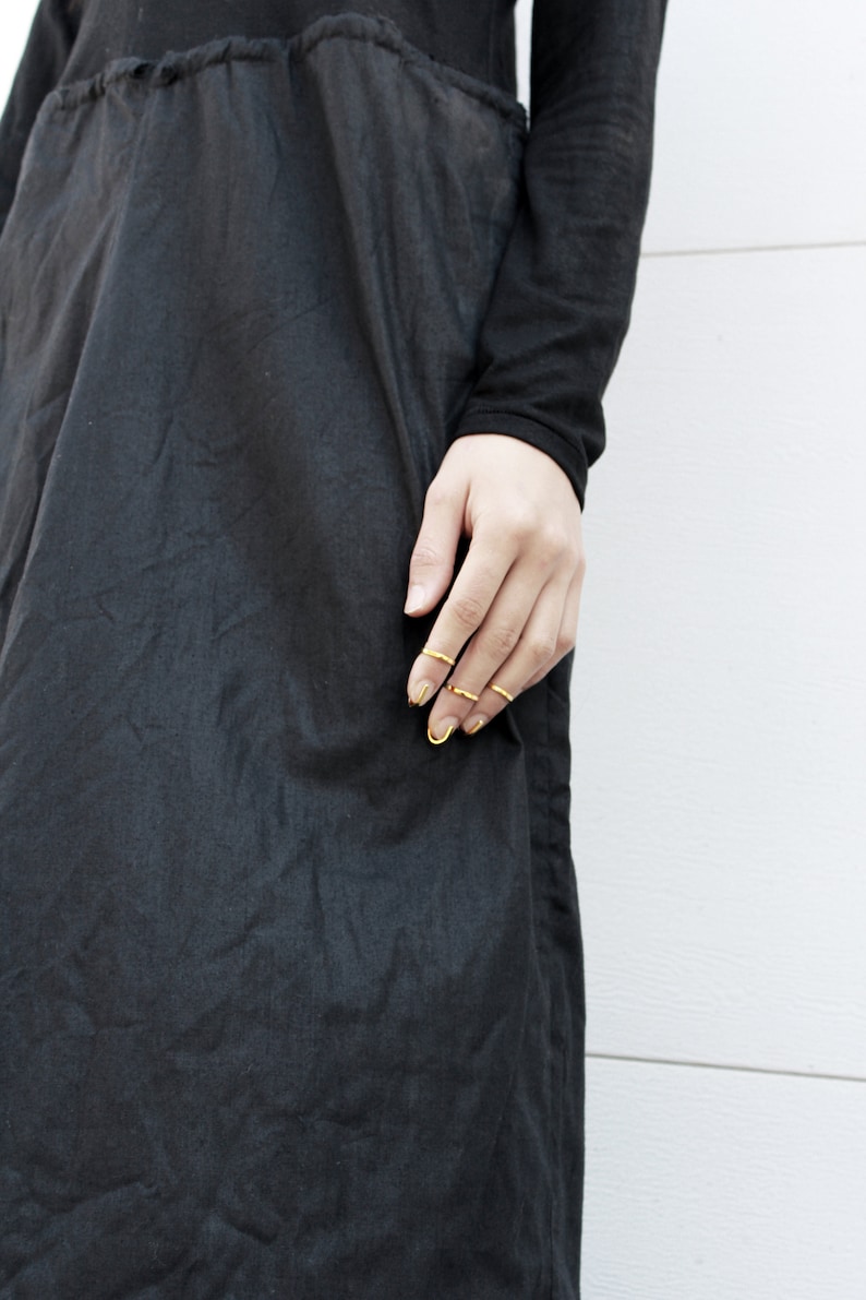 Puede incluir: Top y vestido negros de manga larga. El vestido tiene cintura fruncida y falda fluida. La modelo lleva varios anillos dorados. El fondo es una pared blanca.