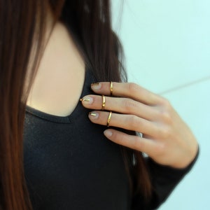 Puede incluir: Primer plano de una mano con anillos dorados y esmalte de u&ntilde;as dorado. La persona lleva una camisa negra de manga larga. El fondo es una pared azul claro.