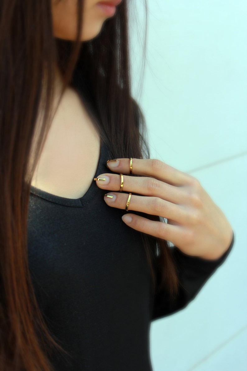 Puede incluir: Primer plano de una mano con anillos dorados y esmalte de u&ntilde;as dorado. La persona lleva una camisa negra de manga larga. El fondo es una pared azul claro.