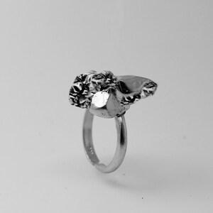 May include: A silver ring with a textured, irregular stone setting. The stone has a mix of smooth and rough surfaces, with a prominent, shiny facet. The ring band is simple and adjustable, creating a unique jewelry piece.