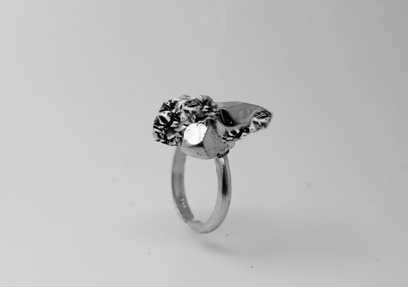 May include: A silver ring with a textured, irregular stone setting. The stone has a mix of smooth and rough surfaces, with a prominent, shiny facet. The ring band is simple and adjustable, creating a unique jewelry piece.