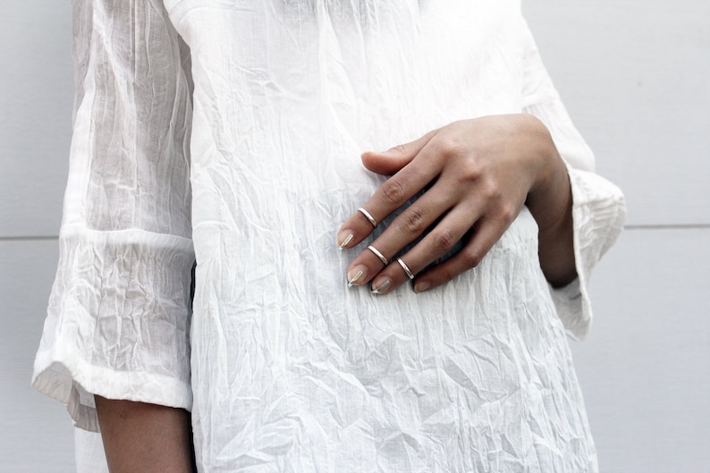 May include: A close-up of a hand wearing several thin silver rings on the fingers. The hand is resting on a white, textured fabric.