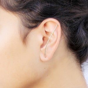 May include: A close-up of a person's ear with a thin gold ear cuff. The cuff is curved and extends from the top of the ear to the lobe.