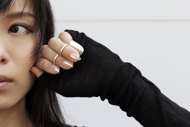 May include: A woman's hand wearing several silver rings on her fingers. The rings are different shapes and sizes, and some are stacked on top of each other. The woman's nails are painted with a silver polish.