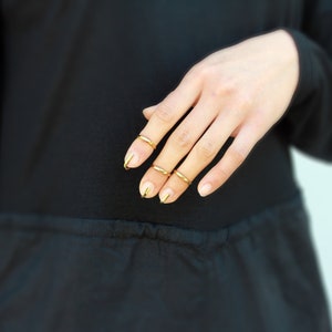 Puede incluir: Una mano con anillos dorados en cuatro dedos y esmalte de u&ntilde;as dorado. La mano se coloca contra una prenda negra, con un fondo azul claro.