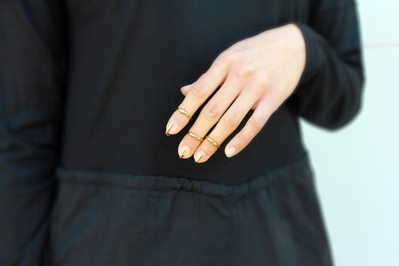 Puede incluir: Una mano con anillos dorados en cuatro dedos y esmalte de u&ntilde;as dorado. La mano se coloca contra una prenda negra, con un fondo azul claro.