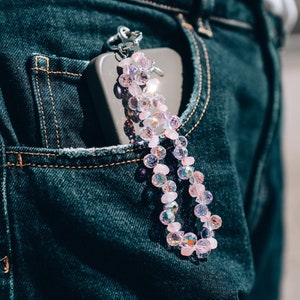 May include: Close-up of a dark blue denim pocket with a smartphone and a beaded keychain. The keychain features pink and iridescent beads, attached to a silver-colored clasp. The denim has visible stitching.