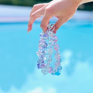 May include: A multi-strand beaded necklace with iridescent blue, pink, and clear beads. The necklace is held by a hand against a blue background.