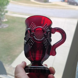 May include: A ruby red glass mug with a decorative handle and base. The mug features an embossed pattern of bows and diamond shapes. The handle is curved and detailed. The mug is held up in front of a window.