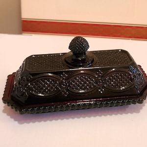 May include: A dark red, rectangular butter dish with a textured lid and base. The lid features a decorative knob, and the dish has an ornate, patterned design. The dish is made of glass and sits on a white surface.