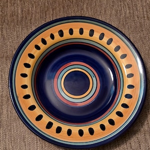 May include: A round, ceramic plate with a vibrant design. The plate features a deep blue center, surrounded by concentric rings of orange, teal, red, and yellow. The outer rim is orange with evenly spaced black dots. A decorative piece for the kitchen.