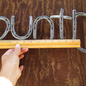 May include: The word "Country" spelled out using horseshoes, with a ruler measuring the length of the word. The horseshoes are silver in color and are nailed to a brown wooden surface.